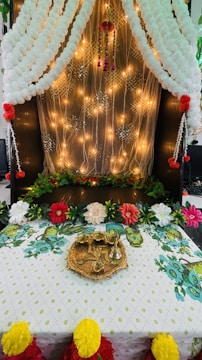 A beautifully decorated area featuring draped white fabric and warm lighting. The background is adorned with glowing string lights and patterns, while the foreground displays an arrangement of colorful artificial flowers and greenery. On the table, there's a decorative metal plate with intricate designs.