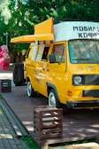 A bright yellow mobile coffee van is parked on a wooden platform surrounded by greenery. The van has an extended awning, and several menu lists or signs are visible on its side. The vehicle is designed to serve as a mobile coffee shop.