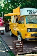 A bright yellow mobile coffee van is parked on a wooden platform surrounded by greenery. The van has an extended awning, and several menu lists or signs are visible on its side. The vehicle is designed to serve as a mobile coffee shop.