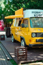 A bright yellow mobile coffee van is parked on a wooden platform surrounded by greenery. The van has an extended awning, and several menu lists or signs are visible on its side. The vehicle is designed to serve as a mobile coffee shop.