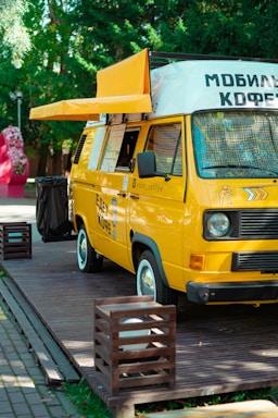 A cheerful barista serving coffee from a vibrant mobile coffee cart parked at a sunny outdoor event.