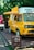 A bright yellow mobile coffee van is parked on a wooden platform surrounded by greenery. The van has an extended awning, and several menu lists or signs are visible on its side. The vehicle is designed to serve as a mobile coffee shop.
