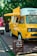 A bright yellow mobile coffee van is parked on a wooden platform surrounded by greenery. The van has an extended awning, and several menu lists or signs are visible on its side. The vehicle is designed to serve as a mobile coffee shop.