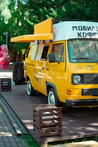 A bright yellow mobile coffee van is parked on a wooden platform surrounded by greenery. The van has an extended awning, and several menu lists or signs are visible on its side. The vehicle is designed to serve as a mobile coffee shop.