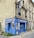 A small embroidery shop with a vibrant blue facade situated on the corner of a street. The windows display designs reminiscent of traditional embroidery, and there is a classic streetlamp mounted on the side of the building. Potted plants add a touch of green to the window ledges above.
