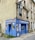 A small embroidery shop with a vibrant blue facade situated on the corner of a street. The windows display designs reminiscent of traditional embroidery, and there is a classic streetlamp mounted on the side of the building. Potted plants add a touch of green to the window ledges above.