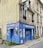 A small embroidery shop with a vibrant blue facade situated on the corner of a street. The windows display designs reminiscent of traditional embroidery, and there is a classic streetlamp mounted on the side of the building. Potted plants add a touch of green to the window ledges above.