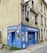 A small embroidery shop with a vibrant blue facade situated on the corner of a street. The windows display designs reminiscent of traditional embroidery, and there is a classic streetlamp mounted on the side of the building. Potted plants add a touch of green to the window ledges above.