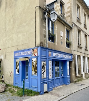 A small embroidery shop with a vibrant blue facade situated on the corner of a street. The windows display designs reminiscent of traditional embroidery, and there is a classic streetlamp mounted on the side of the building. Potted plants add a touch of green to the window ledges above.
