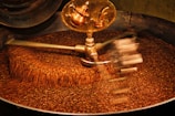 A large batch of coffee beans being roasted inside an industrial machine. The beans are in constant motion due to metallic arms rotating and stirring them. A shiny, copper part of the machine is visible at the top.