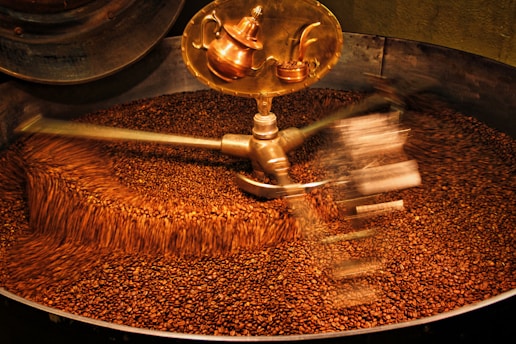 A large batch of coffee beans being roasted inside an industrial machine. The beans are in constant motion due to metallic arms rotating and stirring them. A shiny, copper part of the machine is visible at the top.