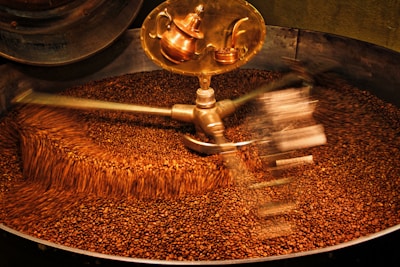A large batch of coffee beans being roasted inside an industrial machine. The beans are in constant motion due to metallic arms rotating and stirring them. A shiny, copper part of the machine is visible at the top.
