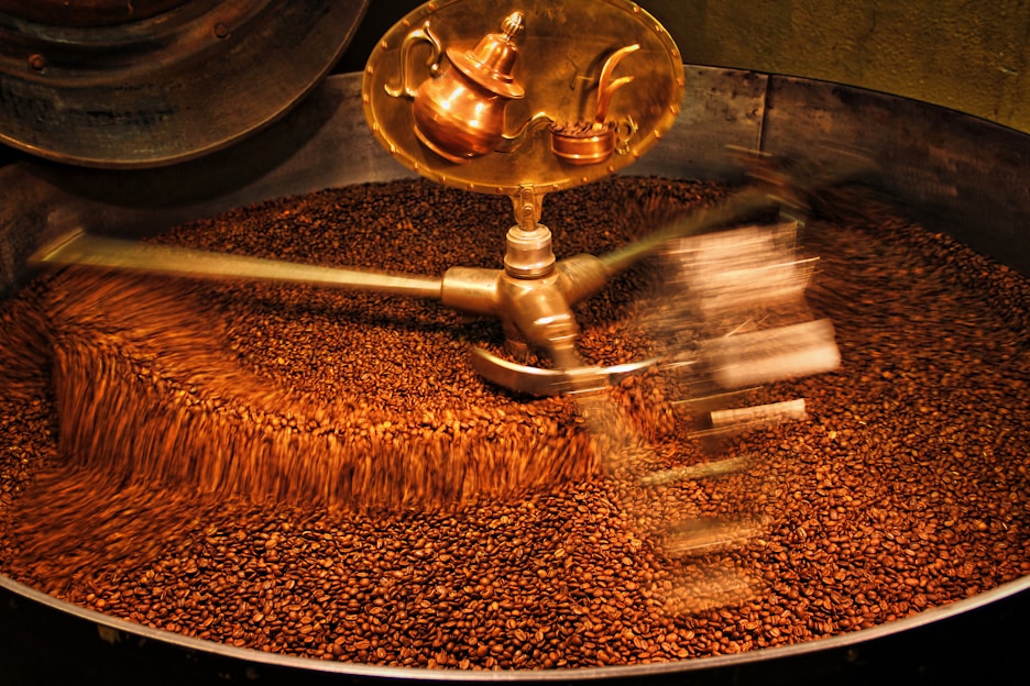 A large batch of coffee beans being roasted inside an industrial machine. The beans are in constant motion due to metallic arms rotating and stirring them. A shiny, copper part of the machine is visible at the top.