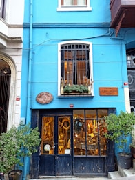 A quaint storefront with a bright blue facade is adorned with a wooden sign and potted plants. The lower level features large windows displaying a variety of antique items inside, including mirrors and furniture. Above, there is a white-paned window with potted plants.