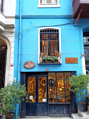 A quaint storefront with a bright blue facade is adorned with a wooden sign and potted plants. The lower level features large windows displaying a variety of antique items inside, including mirrors and furniture. Above, there is a white-paned window with potted plants.