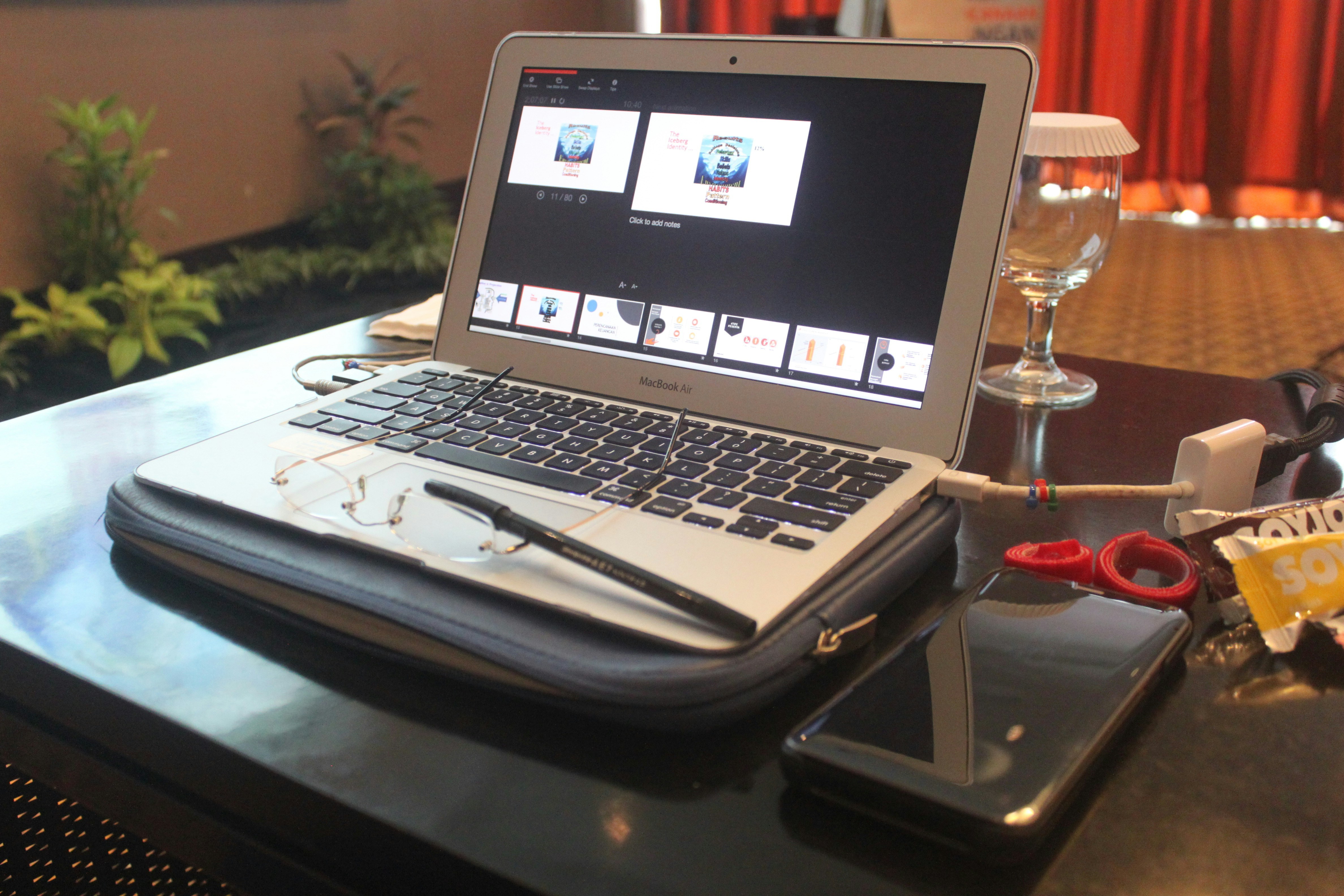 A laptop sits on a table, displaying a presentation slide on the screen. Nearby are a pair of glasses, a pen, a smartphone, and a drink with a glass cover. Various items like cables and small accessories are scattered around. In the background, potted plants and a curtain are visible.