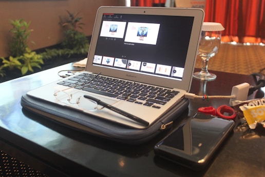A laptop sits on a table, displaying a presentation slide on the screen. Nearby are a pair of glasses, a pen, a smartphone, and a drink with a glass cover. Various items like cables and small accessories are scattered around. In the background, potted plants and a curtain are visible.