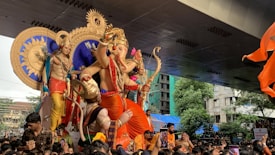 A large idol of a deity with multiple arms and a bow, surrounded by people in a festive procession. The idol is ornately decorated with vibrant orange and red garments, and there is a golden sun motif in the background. A canopy is visible overhead, and colorful flags are waving.
