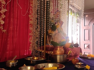 A beautifully decorated altar with a statue of a deity, possibly Ganesh, adorned with garlands and surrounded by various offerings such as fruits, a coconut, and metal dishes. The background features pink drapes and hanging crystal strings, creating a festive and divine ambiance.