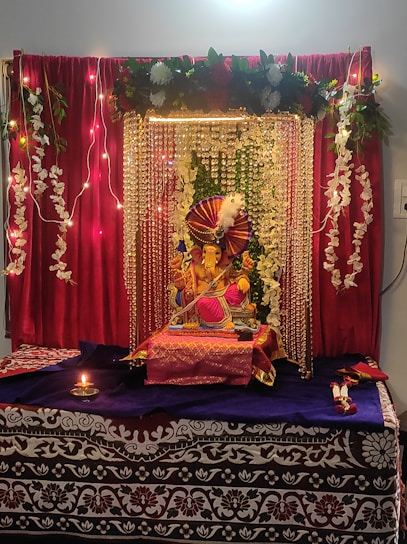 A decorated altar featuring a statue of Ganesha adorned with colorful fabrics, garlands of flowers, and strings of beads. The backdrop includes a rich red curtain with floral arrangements and decorative lights. A small oil lamp is lit in front of the altar, adding to the serene ambiance.