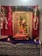 A decorated altar featuring a statue of Ganesha adorned with colorful fabrics, garlands of flowers, and strings of beads. The backdrop includes a rich red curtain with floral arrangements and decorative lights. A small oil lamp is lit in front of the altar, adding to the serene ambiance.