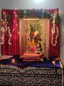 A beautifully decorated altar featuring a Ganesha idol adorned with vibrant decorations. The setting includes red curtains, strings of lights, and garlands of white flowers. The idol is placed on a table covered with purple and red cloth, surrounded by decorative elements. A small oil lamp is lit in front, enhancing the spiritual ambiance.