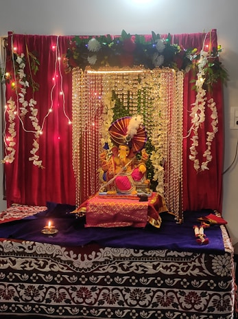 A beautifully decorated altar featuring a Ganesha idol adorned with vibrant decorations. The setting includes red curtains, strings of lights, and garlands of white flowers. The idol is placed on a table covered with purple and red cloth, surrounded by decorative elements. A small oil lamp is lit in front, enhancing the spiritual ambiance.