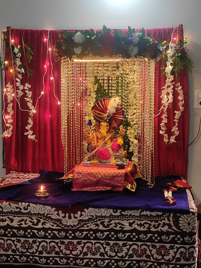 A beautifully decorated altar featuring a Ganesha idol adorned with vibrant decorations. The setting includes red curtains, strings of lights, and garlands of white flowers. The idol is placed on a table covered with purple and red cloth, surrounded by decorative elements. A small oil lamp is lit in front, enhancing the spiritual ambiance.
