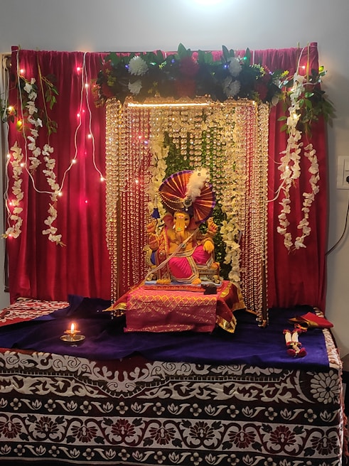 A beautifully decorated altar featuring a Ganesha idol adorned with vibrant decorations. The setting includes red curtains, strings of lights, and garlands of white flowers. The idol is placed on a table covered with purple and red cloth, surrounded by decorative elements. A small oil lamp is lit in front, enhancing the spiritual ambiance.
