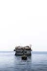 A floating bar and restaurant sits atop calm ocean waters. The structure features thatched roofs and several flags, creating a rustic and inviting appearance. A small boat is anchored in the foreground.