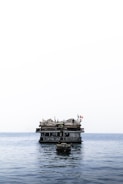 A floating bar and restaurant sits atop calm ocean waters. The structure features thatched roofs and several flags, creating a rustic and inviting appearance. A small boat is anchored in the foreground.
