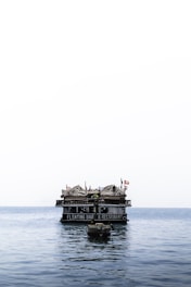 A floating bar and restaurant sits atop calm ocean waters. The structure features thatched roofs and several flags, creating a rustic and inviting appearance. A small boat is anchored in the foreground.