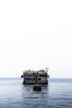 A floating bar and restaurant sits atop calm ocean waters. The structure features thatched roofs and several flags, creating a rustic and inviting appearance. A small boat is anchored in the foreground.