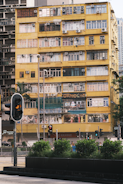 A multi-story urban building with a yellow facade, featuring numerous windows with air conditioning units attached. Several stories are adorned with signs and advertisements. In front of the building, a traffic light is visible, and the street is lined with greenery and some pedestrians.