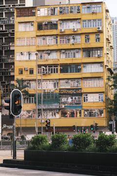A multi-story urban building with a yellow facade, featuring numerous windows with air conditioning units attached. Several stories are adorned with signs and advertisements. In front of the building, a traffic light is visible, and the street is lined with greenery and some pedestrians.