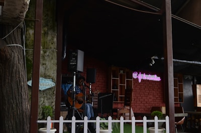 A cozy outdoor cafe setting with a musician playing a guitar on a small stage. The background features a rustic brick wall with a neon sign that reads 'Cafe de L'amour'. The surrounding area is adorned with potted plants and there is a white picket fence.