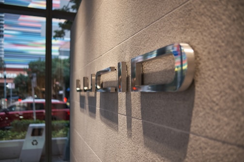 The image shows a modern interior with a close-up focus on metallic letters mounted on a textured wall. The background includes a window view of a street with blurred cars and colorful streaks, possibly from lights or painted structures outside.