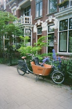 A cargo bicycle is parked on a sidewalk in front of a brick building with large windows, surrounded by greenery and colorful flowers in a residential neighborhood. The architecture features decorative elements and a small balcony.