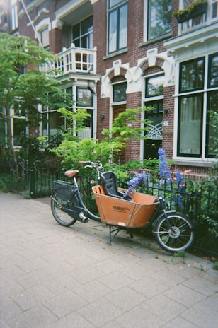 A cargo bicycle is parked on a sidewalk in front of a brick building with large windows, surrounded by greenery and colorful flowers in a residential neighborhood. The architecture features decorative elements and a small balcony.