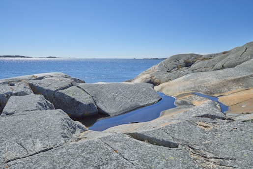 Granite rocks are smoothly contoured by natural erosion, leading down to the gentle, shimmering expanse of a blue sea. Small pools of water have collected in the crevices of the rock formations.