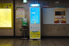 A subway station interior featuring a bright yellow sign with exit information on the left and a green public payphone mounted on a wall. In the middle, there is a blue and white mobile battery rental kiosk. To the right, there is a poster with promotional information in Japanese. The walls are made of textured stones.
