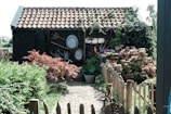 A rustic garden scene features a small wooden shed with a tiled roof. Climbing plants and green foliage partially cover the shed, while an array of gardening tools and metal basins are hung on the side. In the foreground, a variety of flowering shrubs and bushes, including hydrangeas, and a well-maintained path leading through a quaint wooden fence add to the pastoral atmosphere.