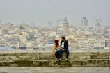 A couple sits on a stone ledge overlooking a sprawling cityscape, with the iconic Galata Tower prominently visible in the background. The city is densely packed with traditional and modern buildings under a hazy sky.