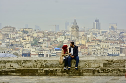 A couple sits on a stone ledge overlooking a sprawling cityscape, with the iconic Galata Tower prominently visible in the background. The city is densely packed with traditional and modern buildings under a hazy sky.