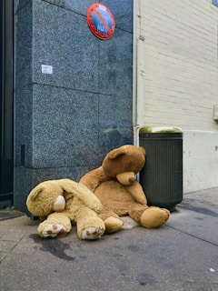 Two large teddy bears are sitting on the sidewalk beside a dark gray granite wall and a black trash can. One bear is upright, leaning against the wall, while the other is slumped over. A red and blue round traffic sign is mounted on the wall above them, and the ground is slightly wet.