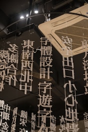 Hanging vertically oriented Asian script characters are suspended from the ceiling against a backdrop of industrial lighting and an air conditioning vent.
