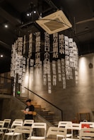 A modern interior space features a decorative installation of hanging white vertical text in Chinese. The environment includes concrete walls and metal elements, with a staircase on the side leading to the upper level. Several white chairs are arranged in rows, suggesting a gathering area. A man dressed in a black shirt stands near the chairs, focused on his phone.