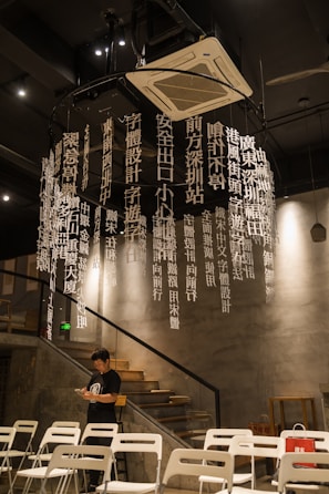 A modern interior space features a decorative installation of hanging white vertical text in Chinese. The environment includes concrete walls and metal elements, with a staircase on the side leading to the upper level. Several white chairs are arranged in rows, suggesting a gathering area. A man dressed in a black shirt stands near the chairs, focused on his phone.