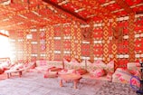A room richly decorated with vibrant, patterned textiles covering the walls, ceiling, and cushions. The prominent colors are bright red, combined with geometric designs in orange, yellow, and black. Several low tables and cushions create a cozy seating area. The overall atmosphere suggests a traditional or cultural setting, possibly inspired by Middle Eastern or North African design.