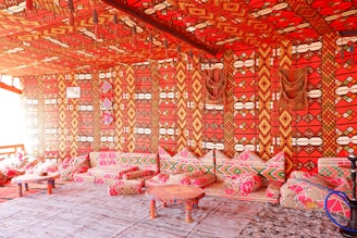 A room richly decorated with vibrant, patterned textiles covering the walls, ceiling, and cushions. The prominent colors are bright red, combined with geometric designs in orange, yellow, and black. Several low tables and cushions create a cozy seating area. The overall atmosphere suggests a traditional or cultural setting, possibly inspired by Middle Eastern or North African design.